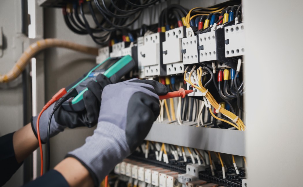Technician performing electrical service upgrades by testing wiring and electrical components inside a commercial electrical panel using a digital multimeter.